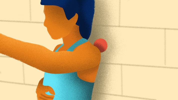 Someone with their arm lifted, leaning on a wall with a ball between the wall and the shoulder to self-massage their frozen shoulder